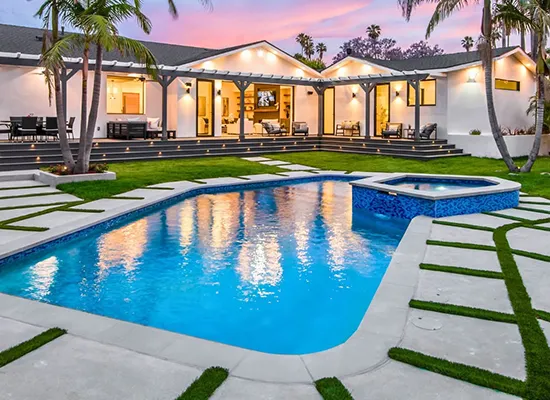 Modern luxury backyard pool with spa, illuminated home, and tropical landscaping at dusk