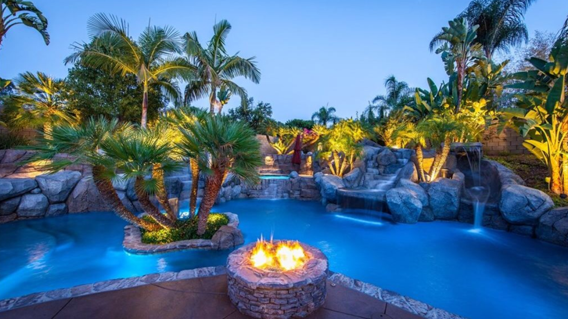 Luxury custom pool with illuminated fire pit, tropical palms, and cascading waterfall features at dusk