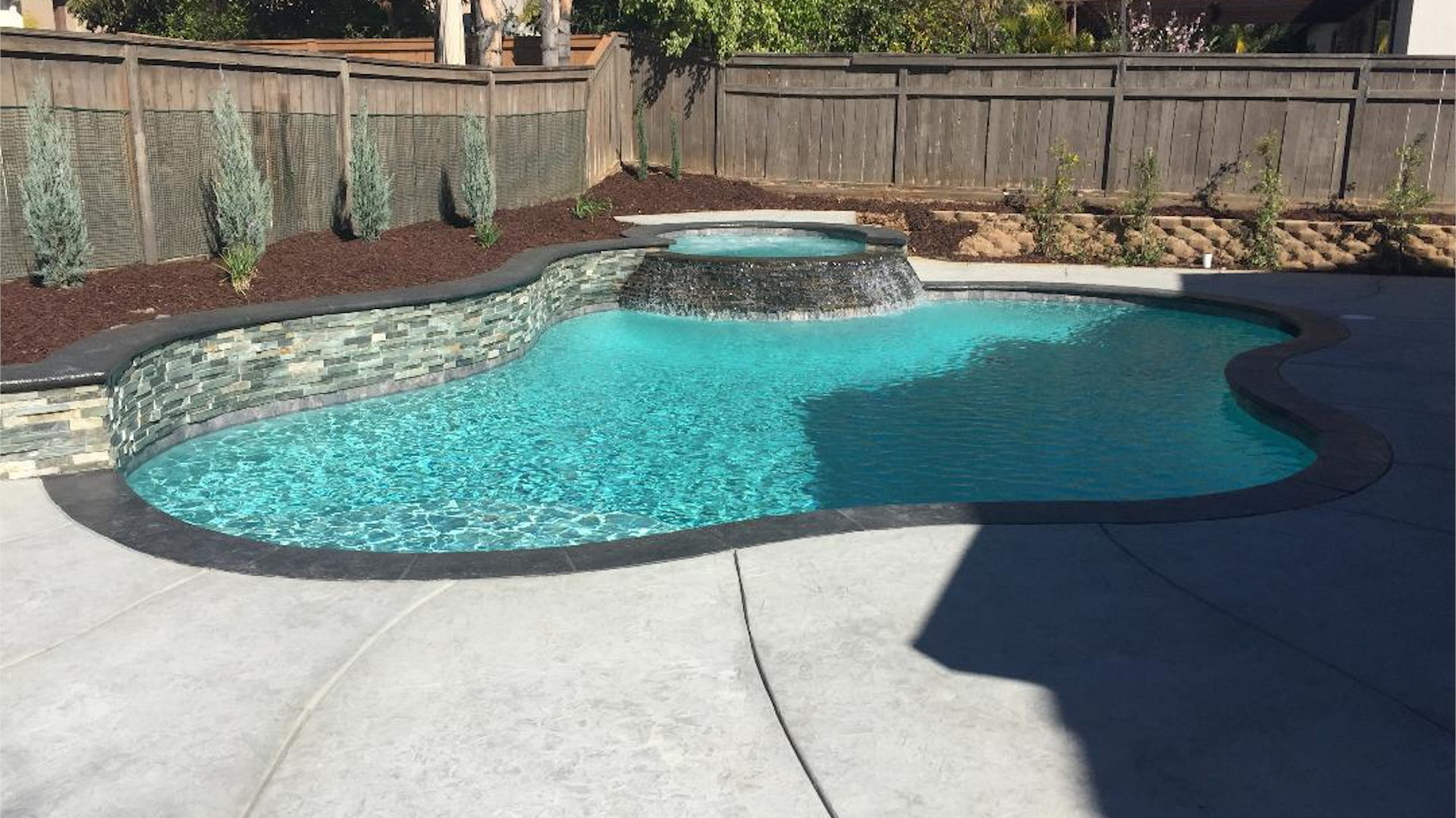 Luxury custom pool with spa waterfall, turquoise water, dark decking, and landscaped backyard